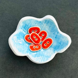 Könnte beinhalten: Eine kleine, weiße Keramikschale mit blauer Glasur und einem welligen Rand. Die Schale enthält sechs rote Keramikknöpfe mit jeweils zwei Löchern.