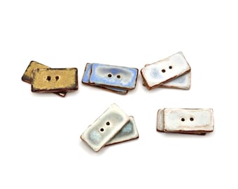 May include: A collection of rectangular ceramic buttons featuring two center holes. The buttons are finished in various glazed colors including metallic gold, light blue, and white, each with a dark brown edge and a smooth, glossy surface texture.