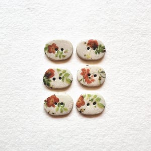 May include: Six oval-shaped buttons with a floral design. Each button features a cream-colored base with a red and green floral pattern. The buttons have two holes for sewing and are arranged in two rows of three.