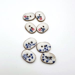 Könnte beinhalten: Sechs ovale Keramikknöpfe mit weißer Basis und braunem Rand. Jeder Knopf ist mit einem handgemalten Blumendesign in Rosa oder Blau verziert. Die Knöpfe haben zwei Löcher zum Annähen, ideal für Bastelprojekte.
