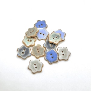 Puede incluir: Un conjunto de doce botones de cerámica con forma de flores de seis pétalos. Cada botón presenta dos orificios centrales y un acabado esmaltado en tonos variados de azul, gris y beige, con bordes marrones visibles en los contornos.