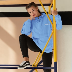 May include: A light blue jacket with the white letters "NLDA" on the chest. The jacket has a zipper and a hood. The person in the image is wearing black pants and black and white sneakers.