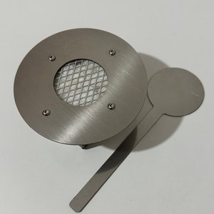 Op de afbeelding: Een zilveren metalen wierookbrander met een ronde basis en een gaas in het midden. Het wordt geleverd met een kleine zilveren metalen lepel.