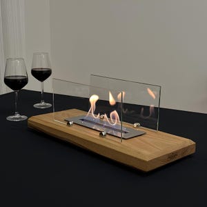 May include: A tabletop fireplace with a wood base and glass panels. The fireplace is lit and has a small flame. The fireplace is on a black surface with two wine glasses.