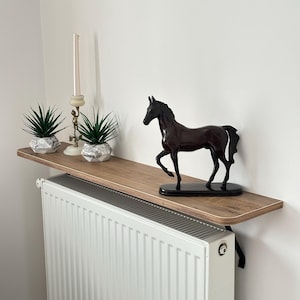 May include: A decorative shelf with a brown wood grain finish, mounted above a white radiator. The shelf displays a dark brown horse figurine, two small potted succulents in geometric pots, and a tall candle holder with a lit candle.