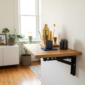 May include: A wooden, slatted, folding table mounted on a white wall. A gold cocktail shaker, jigger, and spoon sit on a wooden tray. A blue cup and a jar of dark beans are also on the table. The table is supported by black metal brackets.