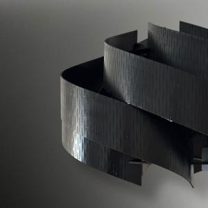 May include: A modern, abstract sculpture featuring layered, curved black panels. The panels have a textured, vertical-lined pattern, creating a sense of depth and movement. The sculpture is set against a neutral gray background.