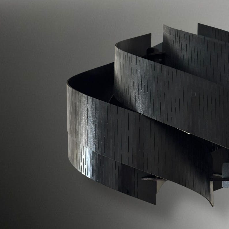 May include: A modern, abstract sculpture featuring layered, curved black panels. The panels have a textured, vertical-lined pattern, creating a sense of depth and movement. The sculpture is set against a neutral gray background.