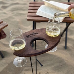 May include: A wooden wine holder with two glass cups and the text "the millies wine n' bar" engraved on the surface. The holder is on a black metal stand and is sitting on a sandy beach.