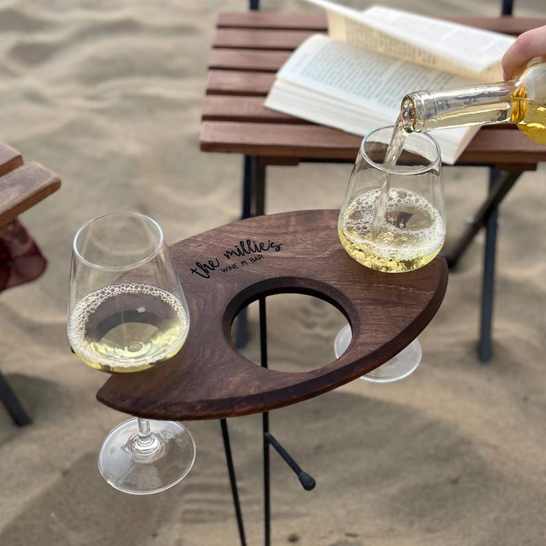 May include: A wooden wine holder with two glass cups and the text "the millies wine n' bar" engraved on the surface. The holder is on a black metal stand and is sitting on a sandy beach.