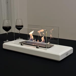 May include: A white tabletop fireplace with a stainless steel burner and glass panels. Flames are visible within the fireplace. Two wine glasses with red wine are in the background. The fireplace has the text "CREEFIRE" on the side.