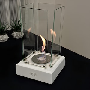 May include: A white, square tabletop fireplace with a flame burning inside a glass enclosure. The base has the word "OFFSHORE" printed on it. The fireplace is surrounded by clear glass panels and has a silver-colored burner.