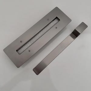 Op de afbeelding: Een zilveren metalen ventilatiekap met een rechthoekige opening en een gaasinsert. Een apart zilveren metalen stuk is inbegrepen voor installatie.