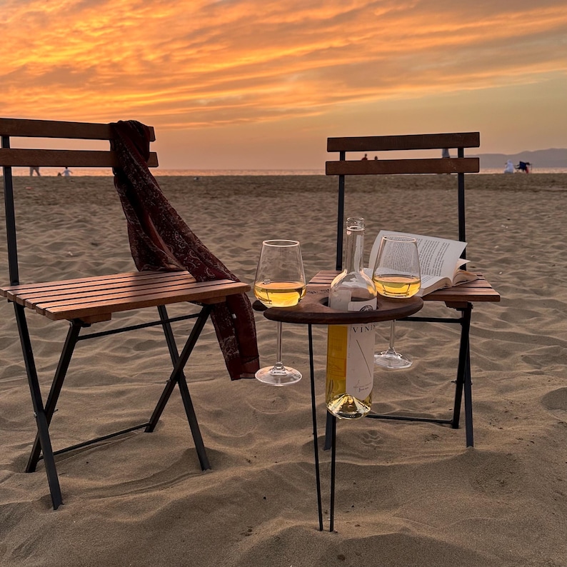 May include: Two folding chairs with a small black metal table between them. The table has a wine bottle holder and two wine glasses. The chairs are on a sandy beach with a sunset in the background.