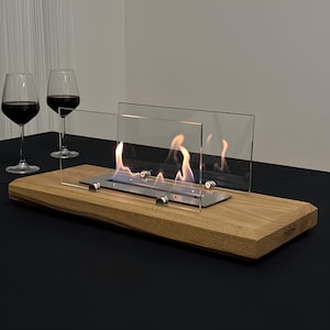 May include: A contemporary tabletop fireplace with a wooden base. Clear glass panels surround the flames, which are visible against a stainless steel burner. Two wine glasses filled with red wine are in the background.