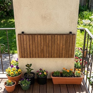 Peut inclure: Une table pliante en bois brun est fixée à un mur beige. Diverses plantes en pot avec des fleurs colorées, dont des pensées et des soucis, sont disposées sur une terrasse en bois. Une balustrade en fer forgé est visible.