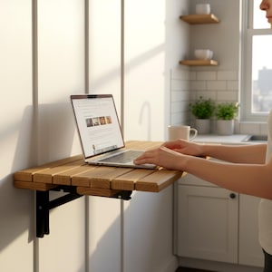 Op de afbeelding: Een houten, aan de muur gemonteerde klaptafel met een laptop erop. De tafel is gemaakt van lichtgekleurd hout en heeft zwarte metalen beugels. Het laptopscherm toont een website. De tafel staat in een keuken.