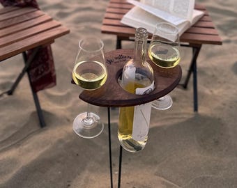 May include: A wooden wine and glass holder with two glasses and a bottle of white wine. The holder is engraved with the text "The Millers" and is sitting on a sandy surface.