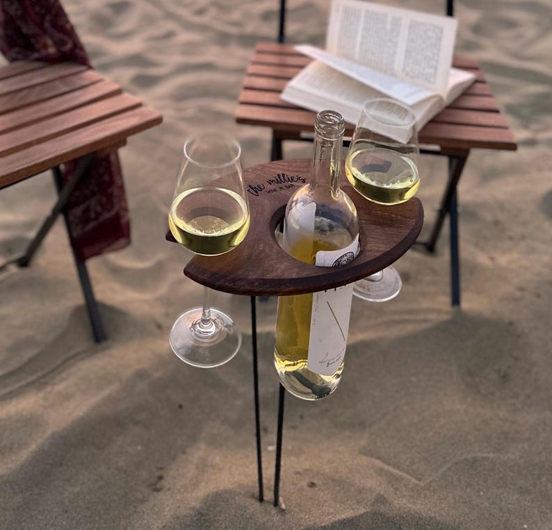 May include: A wooden wine and glass holder with two glasses and a bottle of white wine. The holder is engraved with the text "The Millers" and is sitting on a sandy surface.