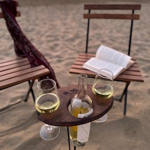 May include: A wooden wine holder with two glasses and a bottle of wine. The holder is engraved with the text "the millie" and "wine in cans". The holder is sitting on a sandy beach with two wooden chairs and a book.