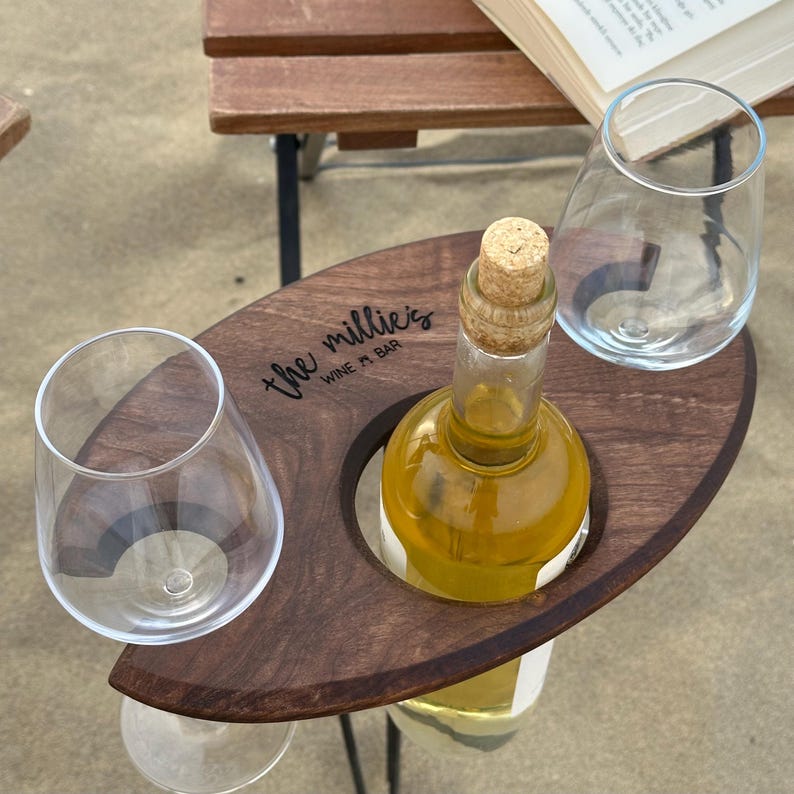 May include: A wooden wine and glass holder with a cutout for a bottle and two glasses. The holder is engraved with the text "the millie's WINE + BAR".