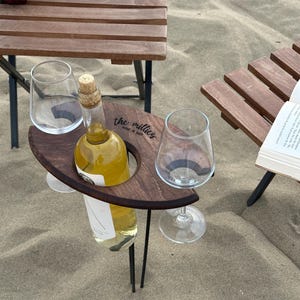 May include: A wooden wine and glass holder with the text "the millies wine & bar" engraved on it. The holder has two glass holders and a bottle holder. The holder is on a black metal stand and is sitting on a sandy beach.