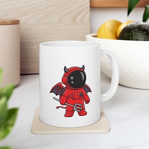 May include: White ceramic mug with a red and black cartoon astronaut with devil horns and a trident. The astronaut is wearing a red spacesuit with a black helmet.