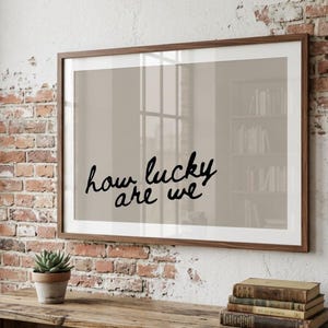 May include: A framed print with the words "how lucky are we" in a handwritten style. The print is displayed on a brick wall above a wooden table. A small potted plant and a stack of books are on the table.