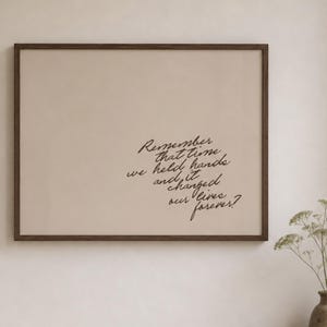 May include: A rectangular wall art piece in a dark brown wooden frame featuring a beige background with black cursive text that reads: Remember that time we held hands and it changed our lives forever? The print is displayed on a plain wall.