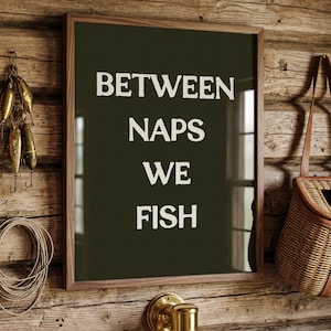 May include: A framed print with the text "BETWEEN NAPS WE FISH" in white letters on a dark green background. The frame is brown, and the print hangs on a wooden wall. Fishing lures and a fishing creel are also visible.