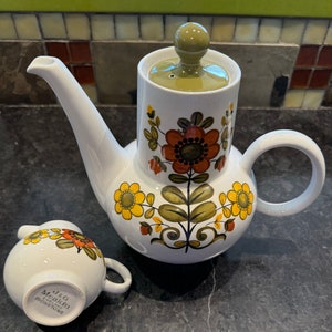 May include: White ceramic teapot and creamer set with a floral design in orange, yellow, and green. The teapot has a curved spout, a handle, and a lid with a green knob. The creamer has a small handle and is marked "J&G Meakin England Ironstone."
