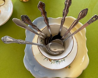 May include: A collection of vintage silverware spoons arranged in a white teacup, resting on a stack of decorative plates. The plates are scalloped, with a yellow and blue color scheme. A yellow creamer with gold trim is visible in the background.