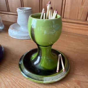 May include: A green ceramic match holder with a black base, holding several matches with pink tips. The holder has a bulbous top and a pedestal base. The base has a decorative rim.