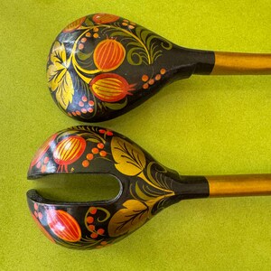 May include: Two wooden spoons with black bowls and gold handles. The bowls are decorated with orange and yellow floral patterns. One spoon has a split bowl design.