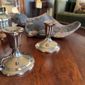 Puede incluir: Dos candelabros de metal plateado pulido con un acabado liso y reflectante. Cada candelabro presenta una base circular ancha que se estrecha hacia arriba en un tallo delgado, rematado con una copa ensanchada para sostener una vela.
