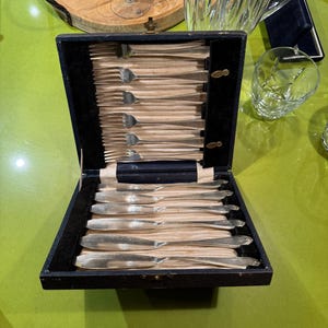 May include: An open, navy blue cutlery box with a set of silver forks and knives. The box is lined with black velvet and contains multiple pieces of silverware. A wooden tray with wine glasses and a crystal pitcher are in the background.