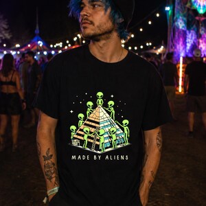 May include: A black short-sleeved t-shirt featuring a graphic print of several green aliens constructing a large pyramid. The design includes white stars in the background and the text "MADE BY ALIENS" printed in white capital letters below the pyramid.