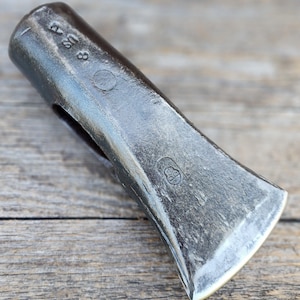 Op de afbeelding: Een donkergrijze metalen bijlkop met een verweerd, getextureerd oppervlak. De zijkant van de bijlkop is voorzien van de gestempelde tekst 8 US en een cirkelvormig merkteken, evenals een klein, duidelijk logostempel bij de scherpe, gebogen snede.