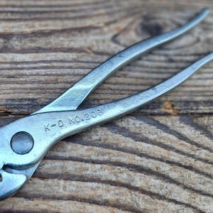May include: A silver K-D No. 203 tool, possibly pliers, with curved handles and a gripping head. The tool is set against a weathered wooden surface, highlighting its industrial design and vintage appeal. The text "K-D No. 203" is visible.
