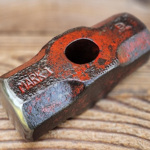 May include: A heavy metal hammer head with a weathered red finish and a central circular opening. The word MARK 1 is embossed on the side of the tool. The surface shows signs of wear, oxidation, and texture, with metallic edges visible at the ends.
