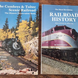 May include: Two books about railways. The first, "The Cumbres & Toltec Scenic Railroad", features a steam train. The second, "Railroad History", shows a maroon and silver train. Both books are on a wooden surface.