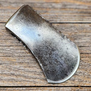 May include: A metal axe head with a dark, weathered, and textured surface. The blade features a curved, sharpened edge that shows signs of wear and use. The poll is rounded, and the eye for the handle is visible on the side of the tool head.