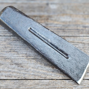 Op de afbeelding: Een donkergrijs metalen wigvormig gereedschap met een getextureerd, gesmeed oppervlak. Het object heeft een taps toelopende, platte punt en een afgerond, stomp uiteinde. Een ondiepe, langwerpige groef is gecentreerd op de bovenzijde van de wig.