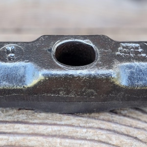 May include: A dark gray metal sledgehammer head featuring a central circular eye for a handle. The surface shows a stamped circular logo on the left side and the text "DROP FORGED" along with "SAFETY" and "6065" embossed on the right side.