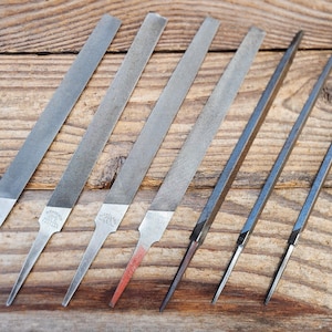 May include: A selection of metal files in different shapes and sizes, displayed on a wooden surface. The files are silver in colour and designed for shaping and smoothing materials. Some files have a red tip.