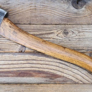 Puede incluir: Un hacha vintage con una hoja de acero pulido y un mango de madera. El mango es de color marrón cálido y la cabeza del hacha es plateada. El hacha descansa sobre una superficie de madera envejecida.