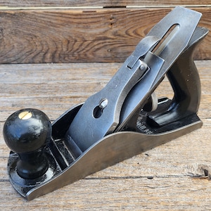 May include: A metal hand plane featuring a black painted body, a polished steel blade assembly, and a rounded black front knob. The tool includes a rear handle and a brass adjustment screw, showcasing a metallic finish and a sturdy construction.