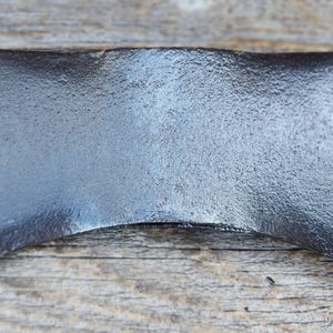 Może przedstawiać: Głowica siekiery dwustronnej wykonana z ciemnej, teksturowanej kutej stali. Metalowa powierzchnia ma szorstkie, matowe czarne wykończenie z polerowanymi, srebrzystymi krawędziami tnącymi po obu stronach. Centralny otwór na trzonek jest niewidoczny.