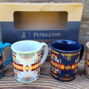 May include: Four ceramic mugs with colorful Native American-inspired designs. The mugs are in shades of blue, white, navy, and tan, with geometric patterns in red, orange, and yellow. A box with the "Pendleton" logo is in the background.
