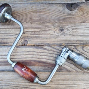 Puede incluir: Taladro manual vintage con mango de madera y marco de metal. El taladro tiene un diseño curvo y un portabrocas para sujetar las brocas. El mango de madera es de color marrón oscuro y el marco de metal es plateado.